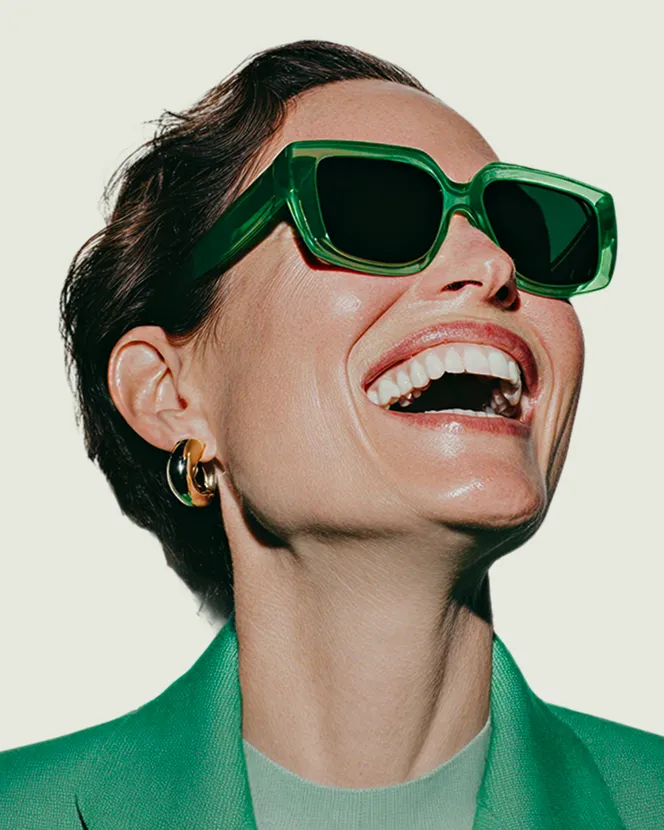 Close-up of a smiling woman wearing large green sunglasses, gold hoop earrings, and a green blazer.